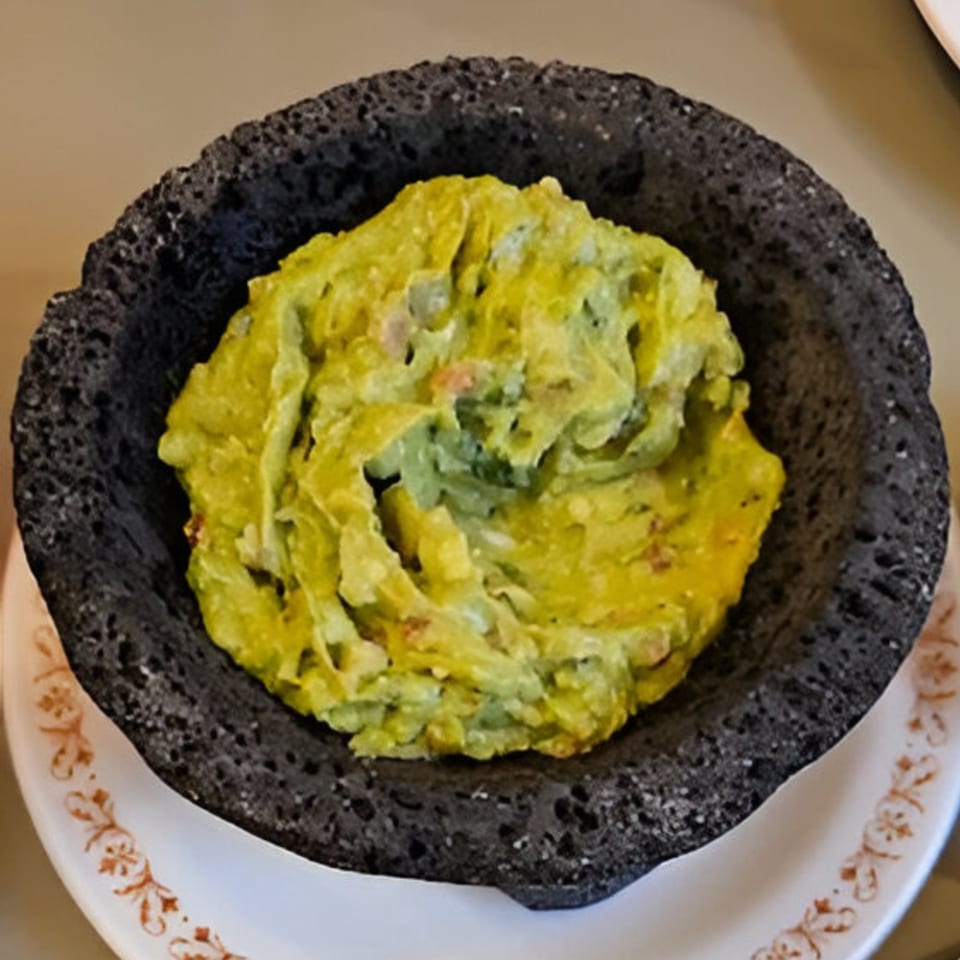 Fresh guacamole in a dark, textured molcajete bowl, served on a white plate with a brown pattern.