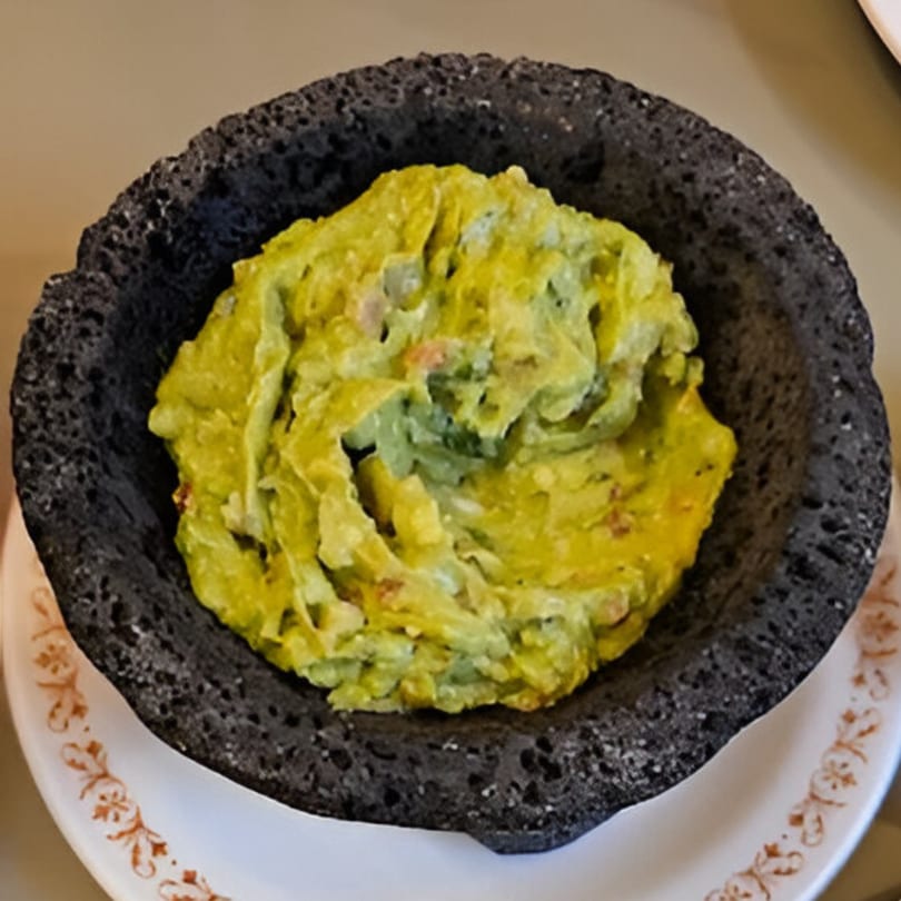 Fresh guacamole in a dark, textured molcajete bowl, served on a white plate with a brown pattern.