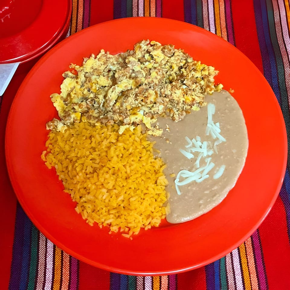 A red plate holds scrambled eggs, yellow rice, and refried beans with shredded cheese on a colorful striped tablecloth.